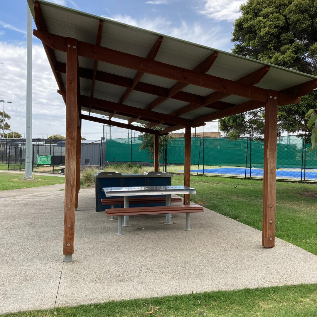 A barbeque and picnic table and chairs are covered by an angled corrugated iron roof. In the background are tennis courts with a blue playing surface.