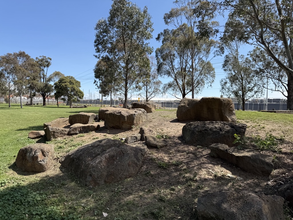 A cluster of large rocks sits on a grassy area surrounded by trees in an open park setting.