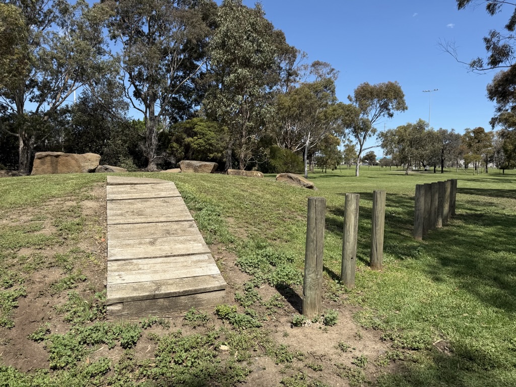 A wooden ramp and upright posts form part of a dog agility course on a grassy area surrounded by trees.