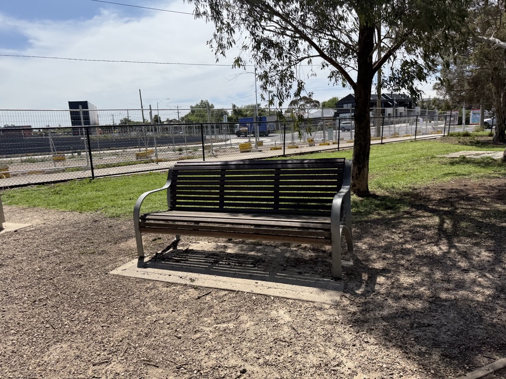 A wooden park bench with metal armrests sits under a tree near a fenced area and construction site.