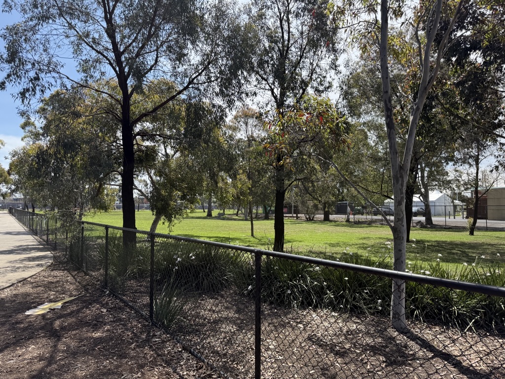 A shaded grassy area with scattered trees is enclosed by a black chain-link fence alongside a paved path.