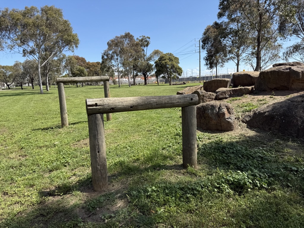 Two wooden hurdle-style posts stand on a grassy area next to large rocks in an open park surrounded by trees.