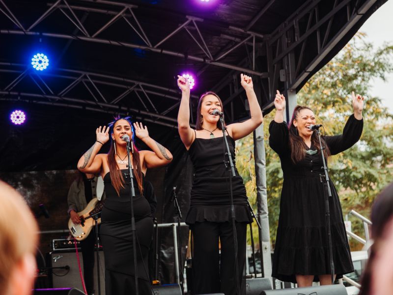 Three performers dressed in black outfits are standing on an outdoor stage under purple lights, raising their hands while singing into microphones, with a guitarist playing in the background.