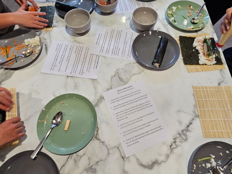 People gathered around a kitchen bench preparing sushi, using bamboo mats to roll seaweed, rice, and fillings with recipe sheets and dishes spread out on the table.