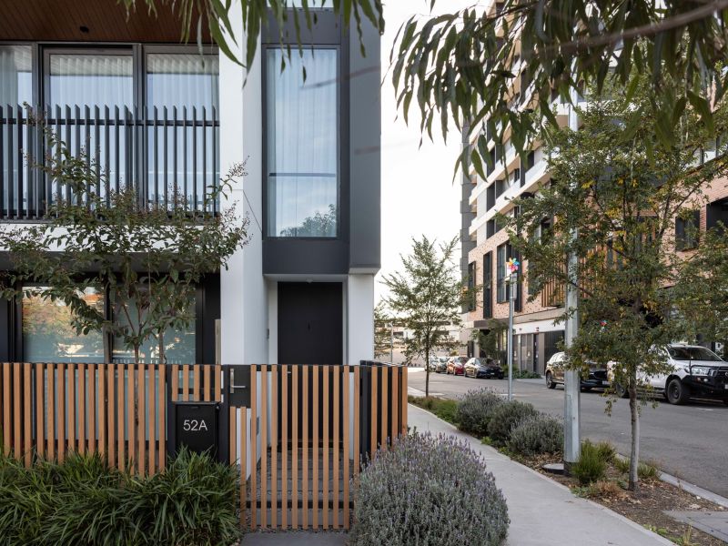 Close view of a modern townhouse with a timber slatted fence and small front garden, set along a tree-lined residential street with parked cars and apartment buildings nearby.