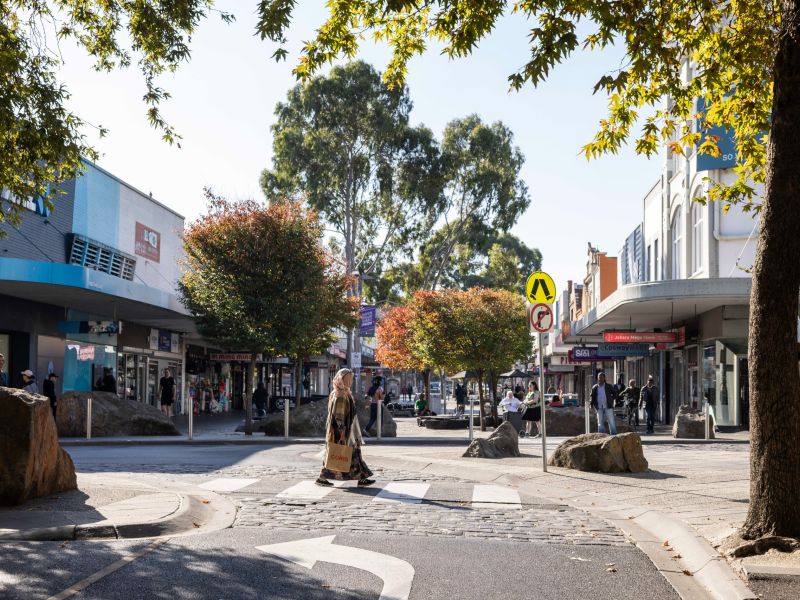 Tree-lined shopping street with people walking across a pedestrian crossing, local shops and signage on both sides, and large rocks and seating in a landscaped streetscape.