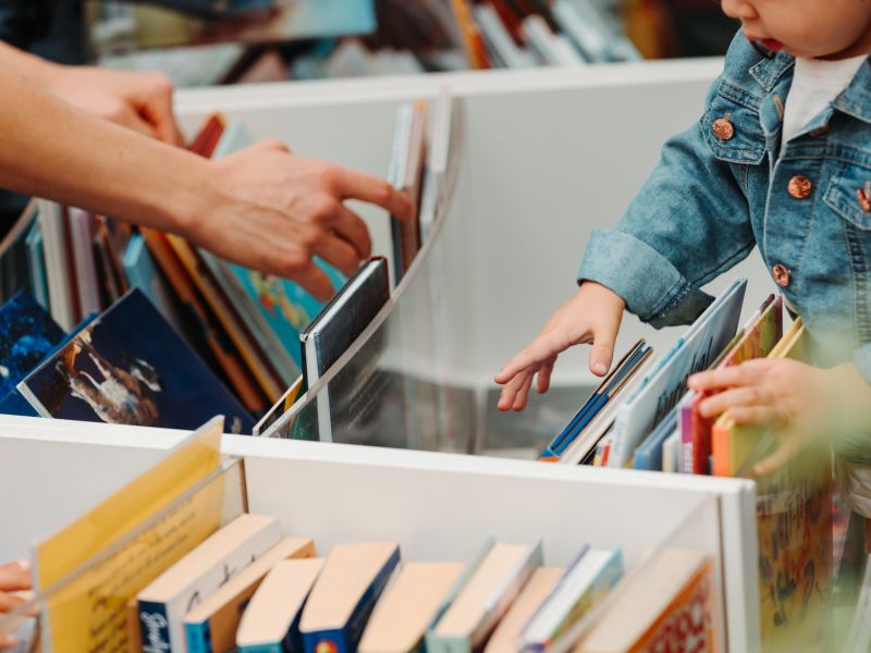 A close-up of hands browsing through a box of colorful books, including children’s titles, with one child reaching toward the books