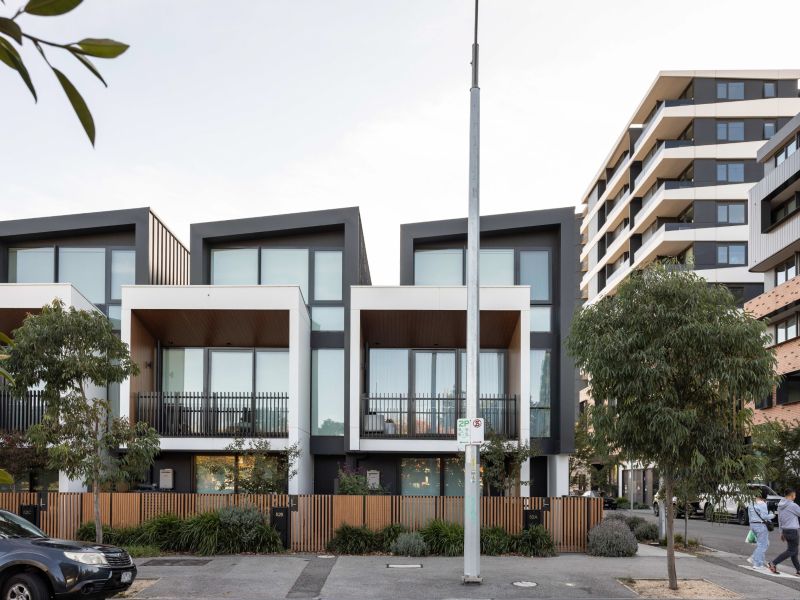 Row of contemporary townhouses with balconies and timber fencing, street trees and a parked car, with a larger apartment building behind and a central light pole in the foreground.