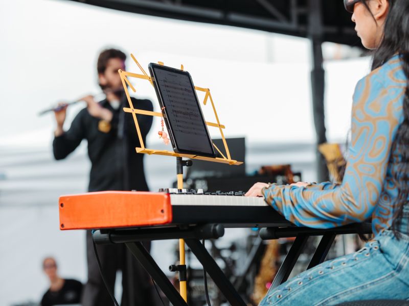 A musician is playing a keyboard with a tablet on a yellow stand displaying sheet music, while another musician in the background plays a flute on an outdoor stage.