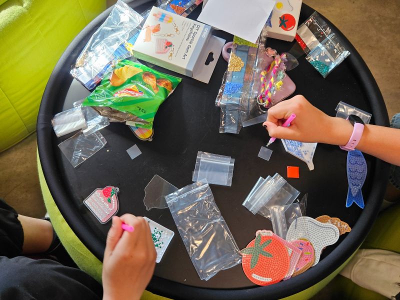People seated around a small table creating bead art crafts, with colourful kits, tools, and materials spread out while placing tiny beads onto shaped templates.