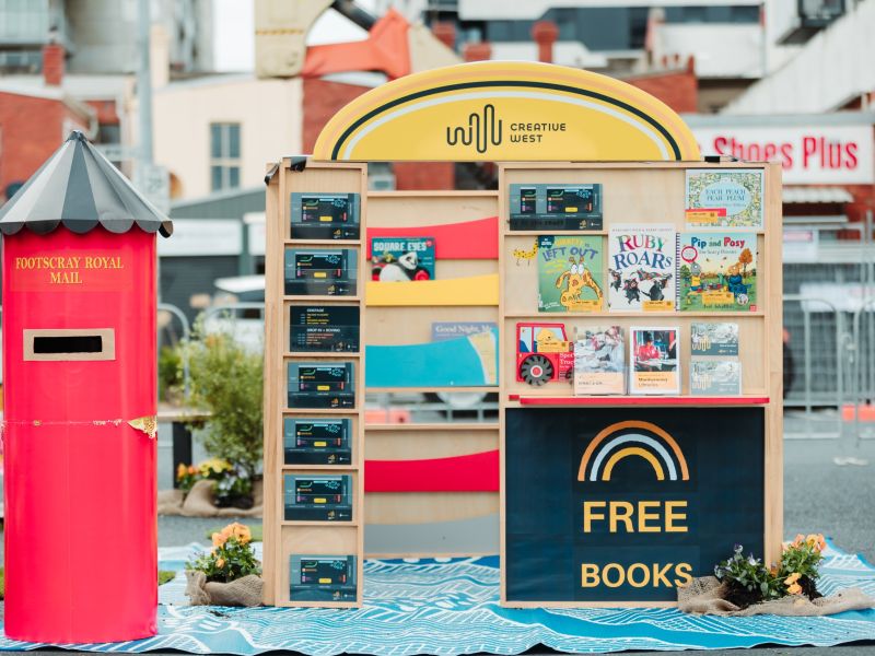A colorful outdoor book display labeled “FREE BOOKS” features children’s books and a bright red “Footscray Royal Mail” mailbox, set against an urban backdrop.
