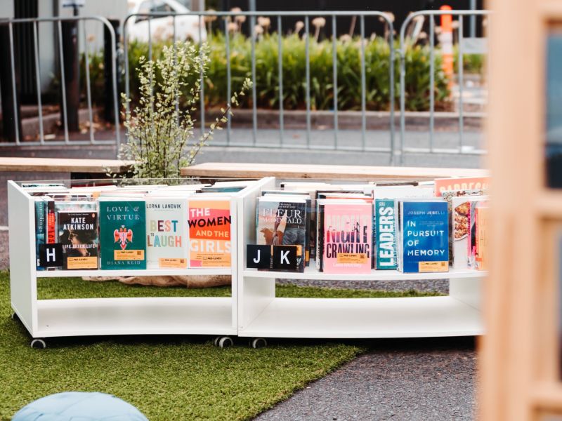 Two white book carts filled with colorful books are displayed outdoors on artificial grass, with titles like Love & Virtue, The Women and the Girls, and In Pursuit of Memory, against a backdrop of a street with fencing and a 20 km/h speed bump sign.