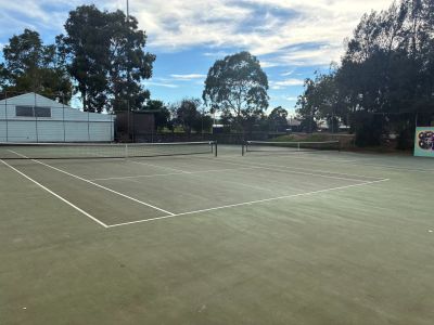 Outdoor tennis courts at Yarraville Oval with marked asphalt surfaces, low fencing and surrounding trees under a partly cloudy sky.