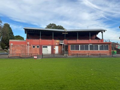 Brick grandstand building with covered seating at Yarraville Oval, viewed from across a green grass playing field under a partly cloudy sky.