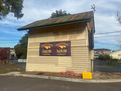 Small timber scorer’s box building at Yarraville Oval with an Eagles banner on the wall, viewed from the surrounding paved area under a partly cloudy sky.
