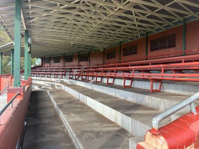 Covered interior of the Yarraville Oval grandstand showing tiered concrete seating with rows of red benches beneath a timber roof structure.